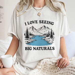 May include: Cream-colored t-shirt with a graphic of mountains, a lake, and trees. The text "I LOVE SEEING BIG NATURALS" is printed above and below the graphic in black. The shirt is a casual, oversized fit.