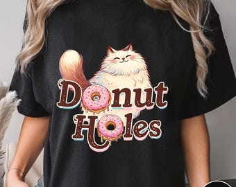 May include: Black t-shirt featuring a cartoon cat holding two pink donuts with sprinkles. The text "Donut Holes" is printed in brown and white. The cat is white and fluffy with a brown tail and ears. The shirt is a comfortable fit.