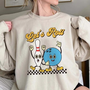 Pode incluir: Uma camisa de moletom bege com uma estampa gráfica de um pino de boliche e uma bola de boliche com rostos de desenhos animados. O texto "Let's Roll" está impresso em amarelo acima do gráfico.