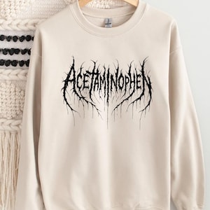 Puede incluir: Una sudadera de color crema con la palabra "ACETAMINOPHEN" en una fuente g&oacute;tica negra y estilizada. El texto tiene un dise&ntilde;o espinoso y org&aacute;nico, que se asemeja a ra&iacute;ces o ramas.