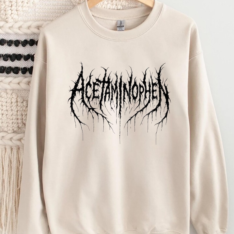 Puede incluir: Una sudadera de color crema con la palabra "ACETAMINOPHEN" en una fuente g&oacute;tica negra y estilizada. El texto tiene un dise&ntilde;o espinoso y org&aacute;nico, que se asemeja a ra&iacute;ces o ramas.