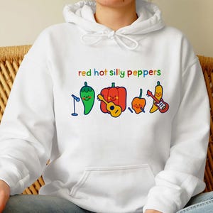 Pu&ograve; includere: Felpa bianca con una grafica rossa, gialla, verde e blu di peperoni con facce e il testo "red hot silly peppers".