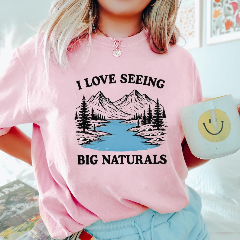 May include: Pink sweatshirt with the text "I LOVE SEEING BIG NATURALS" above a graphic of mountains, trees, and a river. A white mug with a yellow smiley face is held in the hand.