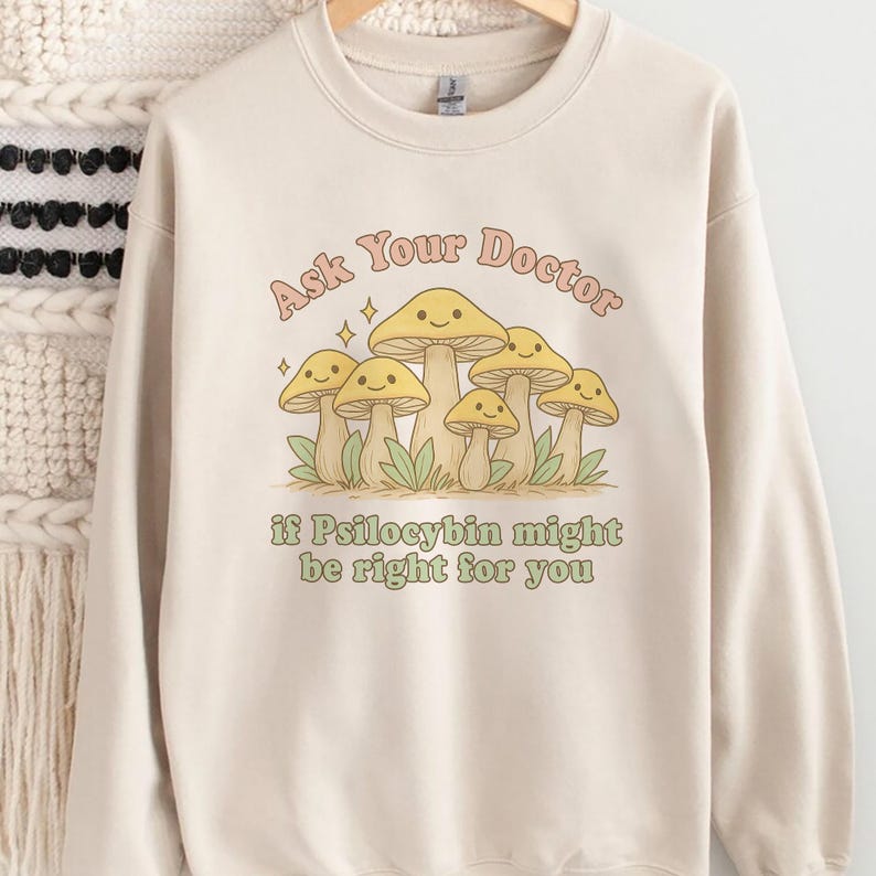 K&ouml;nnte beinhalten: Cremefarbenes Sweatshirt mit einem Pilz-Motiv und dem Text "Ask Your Doctor if Psilocybin might be right for you." Die Grafik zeigt l&auml;chelnde Pilze in Gelb- und Beiget&ouml;nen.