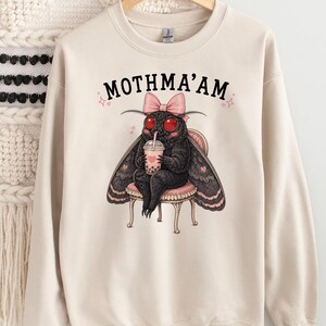 May include: A beige crewneck sweatshirt featuring a graphic of a black moth with red eyes, wearing a pink bow, sitting on a stool, and drinking bubble tea. The text MOTHMA'AM is printed in black above the moth, surrounded by small pink hearts.