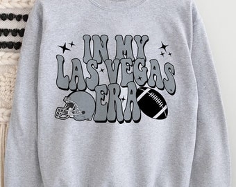 Può includere: Felpa grigia con il testo "In My Las Vegas Era" in un font retrò. Il testo è circondato da stelle e una grafica di un pallone da calcio e un casco.