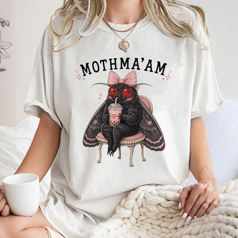May include: An off-white t-shirt featuring a graphic of a black moth with red eyes, wearing a pink bow, sitting on a stool, and holding a boba tea. The text MOTHMA'AM is printed in black above the moth, accompanied by small pink heart and star icons.