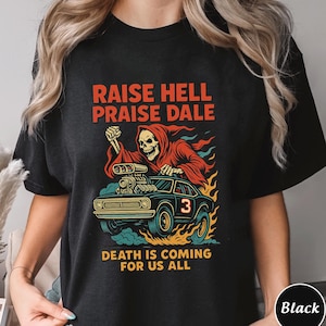 May include: Black t-shirt with a graphic design featuring a grim reaper in a red robe driving a black car with flames. Text on the shirt reads "RAISE HELL PRAISE DALE" and "DEATH IS COMING FOR US ALL".