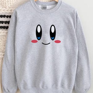 May include: A light gray sweatshirt featuring a cartoon face with large black and white eyes, blue irises, and pink cheeks. The design is centered on the front of the sweatshirt. The sweatshirt is hanging on a wooden hanger.