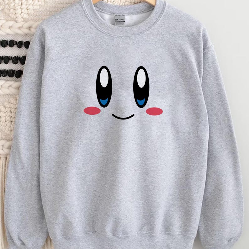 May include: A light gray sweatshirt featuring a cartoon face with large black and white eyes, blue irises, and pink cheeks. The design is centered on the front of the sweatshirt. The sweatshirt is hanging on a wooden hanger.