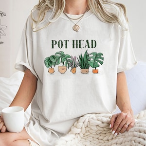 Pode incluir: Camiseta branca com o texto "POT HEAD" em letras verdes e uma fileira de plantas em vasos com rostos de desenhos animados.