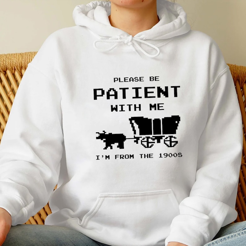 May include: White hooded sweatshirt with black text that reads "Please be patient with me I'm from the 1900s". The text is in a pixelated font and there is a pixelated image of a covered wagon being pulled by an ox.