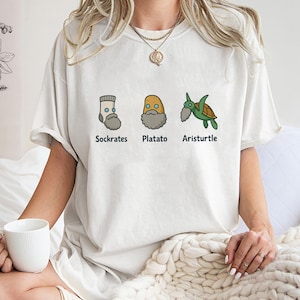 Può includere: T-shirt bianco sporco con tre illustrazioni di cartoni animati e il testo "Sockrates", "Platato" e "Aristurtle". Le illustrazioni raffigurano un calzino, una patata e una tartaruga, ognuno con la barba.