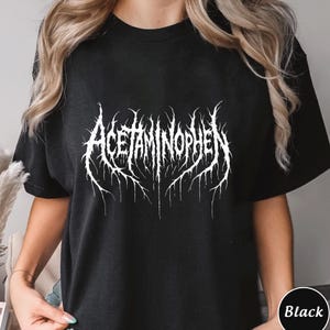 Puede incluir: Camiseta negra con la palabra "ACETAMINOFENO" en blanco, con una fuente de estilo g&oacute;tico. El texto tiene una apariencia desgastada y texturizada, con bordes afilados y puntiagudos. La camiseta es de color negro liso.