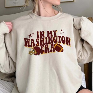 May include: A beige sweatshirt with a maroon graphic that says "In My Washington Era" with a football and helmet.