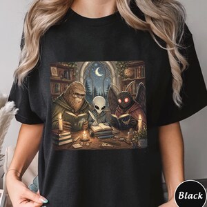 May include: Black t-shirt featuring a fantasy illustration of a Bigfoot, an alien, and a moth-like creature reading books around a table. The scene is set in a library with a moonlit window. The design includes candles and stacks of books.