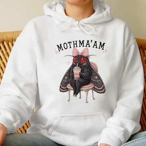 May include: A white hooded sweatshirt featuring a graphic of a black moth with red eyes, wearing a pink bow, sitting on a small stool, and holding a boba tea. The text above the moth reads MOTHMA'AM in black capital letters with small pink stars.