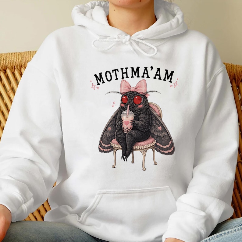 May include: A white hooded sweatshirt featuring a graphic of a black moth with red eyes, wearing a pink bow, sitting on a small stool, and holding a boba tea. The text above the moth reads MOTHMA'AM in black capital letters with small pink stars.