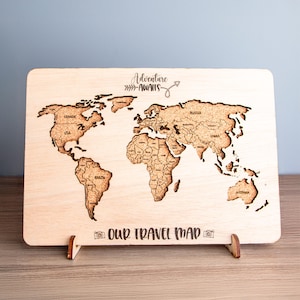 May include: A wooden world map with country outlines and the text "Adventure Awaits" and "Our Travel Map". The map is displayed on a wooden stand. The map is made of light-colored wood.