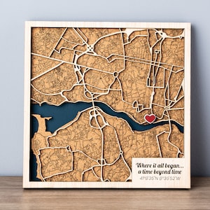May include: Wooden map art featuring a detailed city map with a heart-shaped marker. The map is laser-cut with a brown finish, set against a dark blue background. Text at the bottom reads, "Where it all began..." with coordinates.