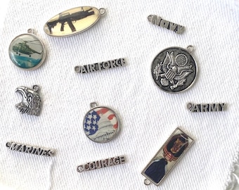 May include: A collection of silver-toned military-themed charms and pendants. The charms include an eagle head, a helicopter, a gun, and the words "Navy", "Airforce", "Marines", "Army", and "Courage". Some charms feature patriotic imagery.