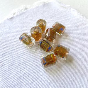 May include: A collection of seven cylindrical glass beads featuring a clear outer layer and an internal swirling pattern of amber, yellow, red, and blue colors. Each bead has a central hole running through the middle of its length.