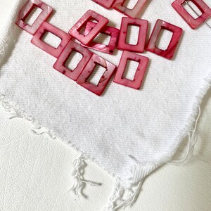 May include: A collection of pink square beads with a rectangular opening, scattered on a white cloth. The beads have a pearlescent sheen and are likely made of shell or a similar material. A silver-colored wire is also present.