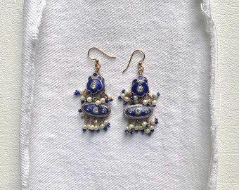 May include: A pair of ornate dangle earrings with gold-colored hooks. The earrings feature a blue enamel design with white and clear accents, and small white beads. The earrings are displayed on a white, textured fabric.