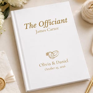 May include: A white book with gold lettering that reads "The Officiant" and "James Carter." Below, it says "Olivia & Daniel" and "October 25, 2026." A gold pen, a ring box with rings, and flowers are in the background.