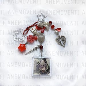 Peut inclure: Un porte-clés en acrylique transparent avec divers breloques. Les breloques comprennent une étoile, un cœur avec des ailes, une étoile rouge, une rose, une seringue et un petit carré avec une illustration de personnage. Le porte-clés a des perles rouges et blanches.