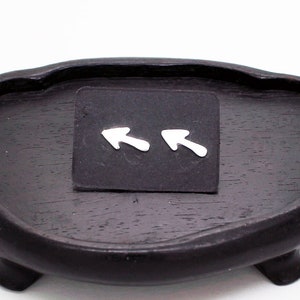 Könnte beinhalten: Eine dunkelbraune, ovale Holzschale mit drei kleinen Füßen. Auf der Schale befindet sich ein kleines, schwarzes Quadrat mit zwei silbernen Pfeilohrringen. Die Ohrringe zeigen in entgegengesetzte Richtungen.