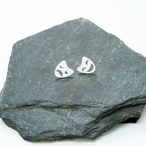 May include: A pair of silver stud earrings with an abstract design. Each earring features cut-out shapes. The earrings are presented on a dark grey, textured surface, providing a contrast with the bright silver of the earrings.