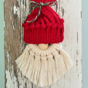 Op de afbeelding: Een rood en wit gehaakte gnome-versiering met een houten kraal neus. De gnome draagt ​​een rode gebreide muts met een groen en bruin lint.