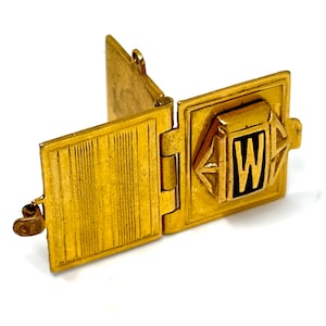 May include: A gold-toned metal hinged locket with three rectangular panels. One panel features a raised central square element with a black letter W set against a gold background. The adjacent panel displays a vertical linear texture pattern.