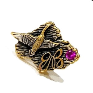 Puede incluir: Broche dorado con un diseño de ave en vuelo. El broche tiene un fondo texturizado, las letras "gB" y una pequeña gema redonda de color magenta.