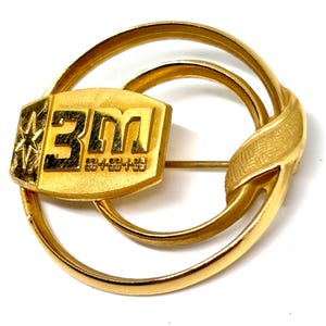 May include: A gold-toned pin featuring the "3M" logo within a rectangular frame, set against a backdrop of intertwined circular and ribbon-like elements. The pin has a classic design with a pin back.