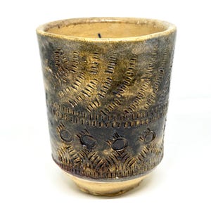 May include: A handmade ceramic tumbler with a textured surface and a unique design. The cup features a dark brown and tan color scheme with etched patterns. The base and rim are lighter in color, while the body is darker.