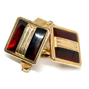 May include: A pair of gold-toned cufflinks with rectangular faces. Each cufflink features a dark red and black enamel design with three gold stripes. The cufflinks are set against a white background.