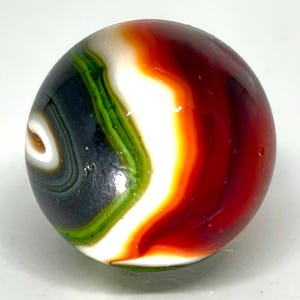 May include: A spherical glass marble featuring swirling bands of color including deep red, bright orange, white, vibrant green, and dark charcoal gray. The surface is smooth and reflective, showcasing a complex, layered pattern of opaque glass.