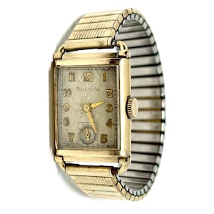 Könnte beinhalten: Eine Vintage-Bulova-Armbanduhr mit einem rechteckigen, goldfarbenen Gehäuse und einem cremefarbenen Zifferblatt. Die Uhr verfügt über goldene Zeiger und Ziffern sowie ein dehnbares, goldfarbenes Armband. Das Zifferblatt zeigt den Markennamen Bulova.