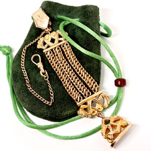 May include: A gold-toned multi-strand chain fob with ornate metalwork at both ends, featuring a decorative seal at the base. The piece includes a small attached chain with a clasp and is displayed against a dark green fabric pouch with green cords.