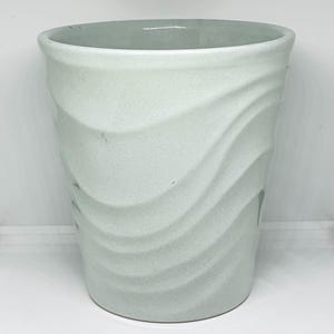 May include: A cylindrical ceramic vessel featuring a light mint green glaze with a subtle speckled texture. The exterior surface is defined by prominent, sweeping diagonal ridges that create a wavy, undulating pattern around the entire body.