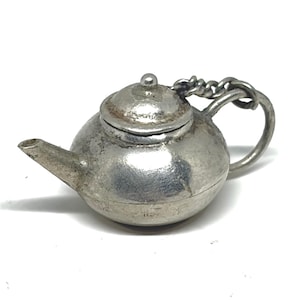 Könnte beinhalten: Ein kleiner, silberfarbener Metallanhänger in Form einer Miniatur-Teekanne. Der Anhänger verfügt über einen abgerundeten Körper, einen kurzen Ausguss, einen gewölbten Deckel mit einem kleinen Knauf und einen gebogenen Griff mit einem Metallring.