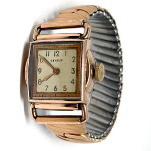 Könnte beinhalten: Vintage Geneva Armbanduhr in Goldoptik mit rechteckigem Zifferblatt. Das cremefarbene Zifferblatt hat schwarze Ziffern. Das Armband besteht aus goldfarbenem Metall und einem dehnbaren silberfarbenen Band. Das Zifferblatt misst ca. 2 cm.