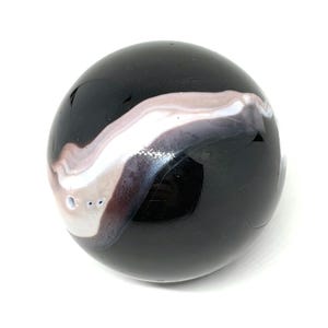 May include: A smooth, spherical black stone bead featuring a prominent, swirling band of white and pale lavender agate-like banding that wraps around the surface. The polished object has a small, circular hole drilled through one side.
