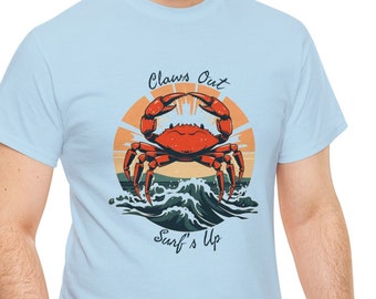 May include: A light blue short-sleeve t-shirt featuring a graphic of a red crab riding a wave in front of a sunset. The text "Claws Out" is printed above the crab, and the text "Surf's Up" is printed below the wave in a dark blue font.