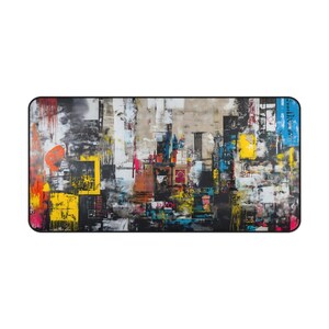 May include: Rectangular desk mat with an abstract cityscape design. The artwork features a collage of colors including black, white, yellow, orange, and blue. The mat has a black border and a smooth surface.