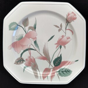 May include: An octagonal, off-white plate with a floral design. The plate features pink roses, green leaves, and brown stems. The plate has a thin, dark blue border. A decorative piece for the dining table.