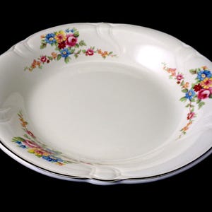 May include: White ceramic serving bowl with a scalloped edge and floral design. The bowl has a silver trim and is decorated with colorful flowers in pink, blue, yellow, and red. The bowl is on a black background.