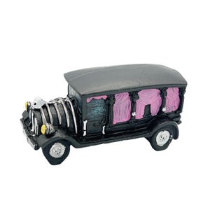 Puede incluir: Un coche fúnebre negro en miniatura con cortinas rosas en las ventanas. El vehículo de estilo vintage tiene un techo gris, faros amarillos y detalles plateados. El coche fúnebre tiene cuatro ruedas y está sobre un fondo blanco.